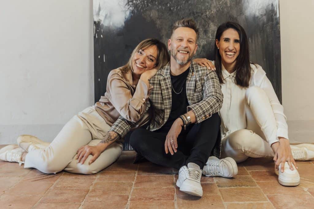 Tres personas sentadas en el suelo en un interior, sonriendo. Las dos mujeres de cada lado están apoyadas contra el hombre que está en el medio. Están vestidas de manera informal.