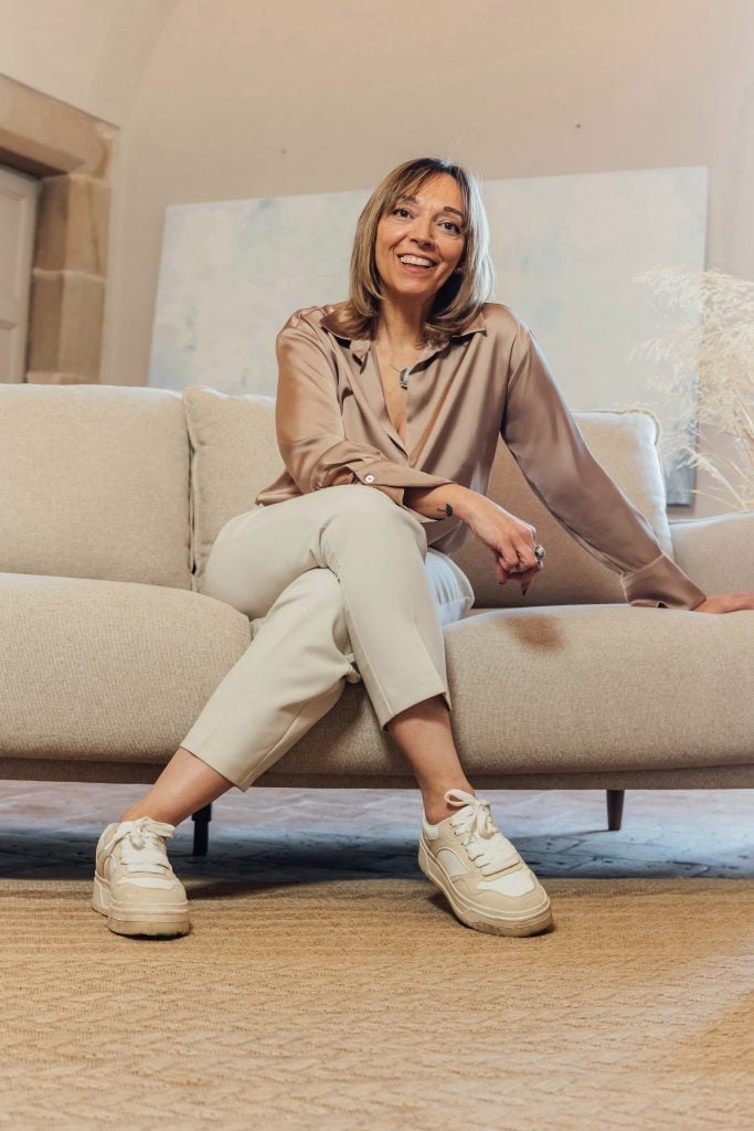 Mujer sentada en un sofá beige, con blusa marrón claro, pantalón blanco y zapatillas blancas. Sonríe y apoya el brazo en el respaldo sobre un fondo neutro.