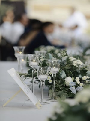 Mesa con centro de mesa con flores, copas de vino y un menú sobre un mantel blanco. Personas en el fondo borroso.