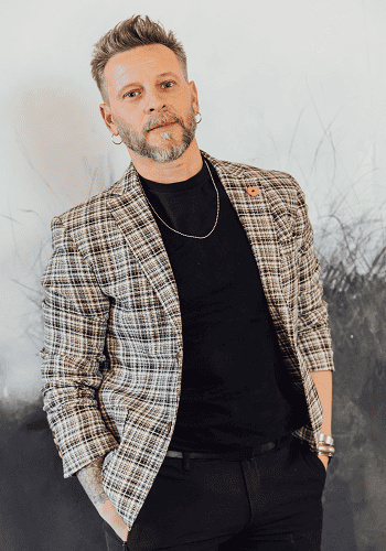 Una persona con barba y pendientes, vestida con una chaqueta a cuadros y una camisa negra, se encuentra sobre un fondo gris.