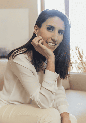 Una mujer de cabello largo y oscuro, vestida con una blusa blanca, está sentada en un sofá, sonriendo con la barbilla apoyada en la mano.