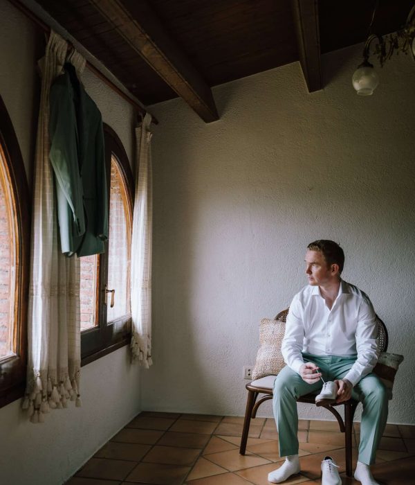 Un hombre con camisa blanca y pantalones verde claro está sentado en una silla, mirando por la ventana de una habitación con vigas de madera en el techo. Una chaqueta verde cuelga junto a la ventana.