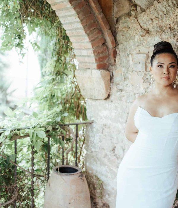 Una mujer con un vestido blanco sin tirantes se apoya contra una pared de piedra con vegetación a su alrededor.