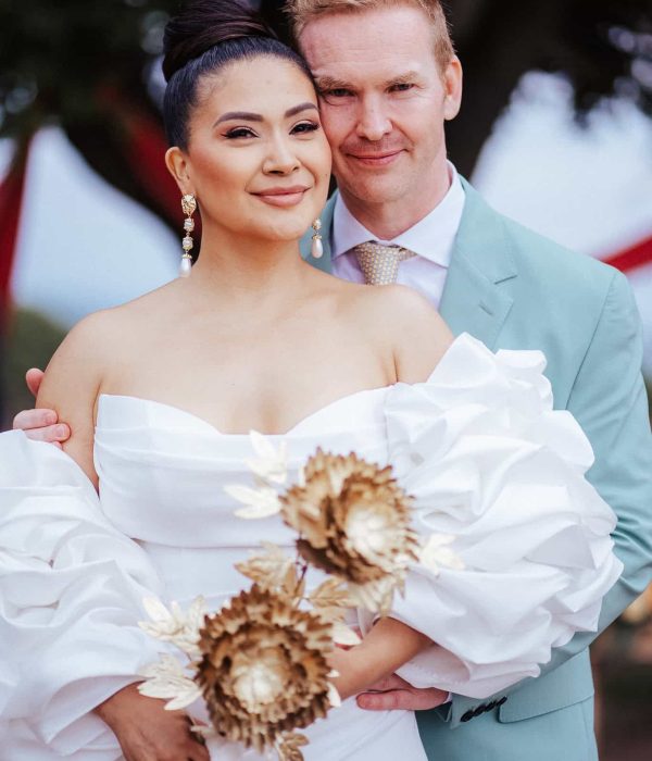 Una pareja con traje de boda posa junta; la mujer sostiene flores. Ella lleva un vestido blanco; él lleva un traje azul claro.