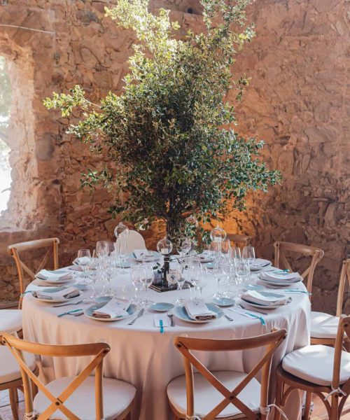 Una mesa redonda preparada para comer con mantel y servilletas blancas, rodeada de sillas de madera. Un gran centro de mesa con plantas verdes se encuentra en el centro de la mesa, en una habitación rústica de piedra.