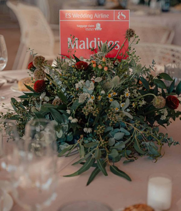 Un centro de mesa con un arreglo floral verde y un cartel rojo con la palabra "Maldivas". Alrededor hay copas de vino, platos y panecillos sobre un mantel blanco.