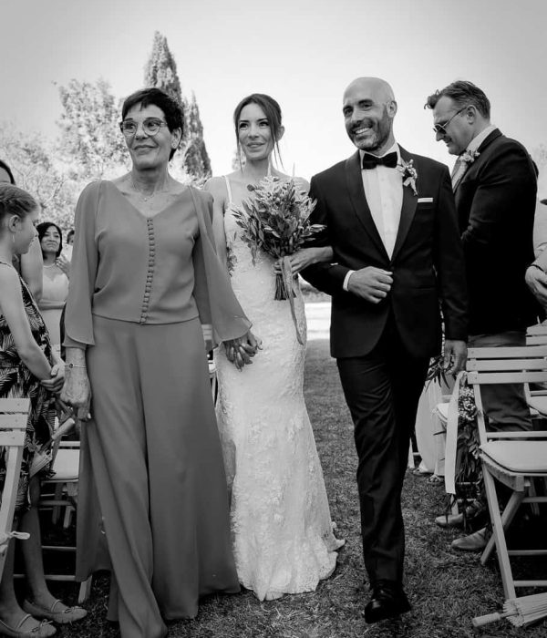 Una novia con un vestido blanco camina hacia el altar con una mujer mayor y un hombre con traje. Están al aire libre, rodeados de invitados sentados.