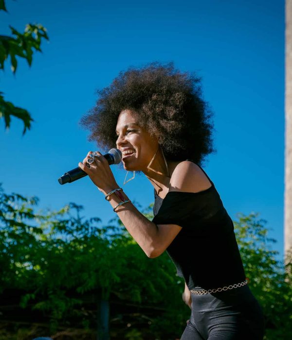 Persona con cabello rizado cantando en un micrófono al aire libre cerca de una torre de piedra, rodeada de vegetación bajo un cielo azul claro.