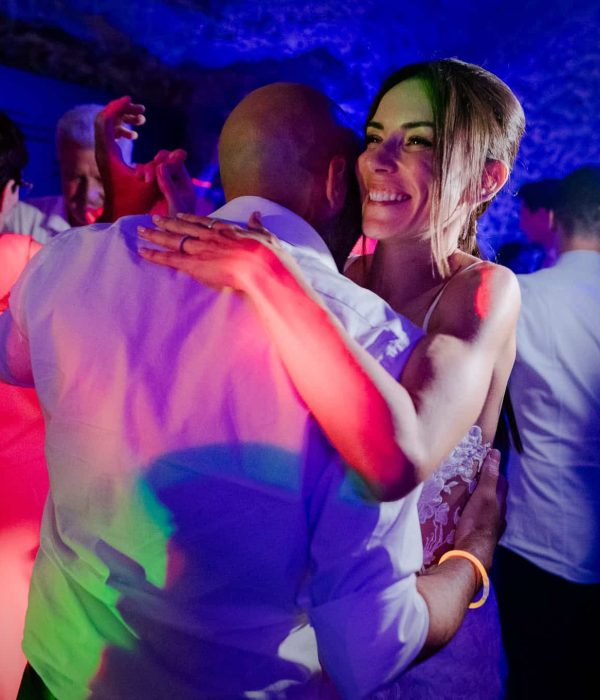Una pareja baila muy cerca en una sala poco iluminada, rodeada de gente. Las luces multicolores crean un ambiente festivo.