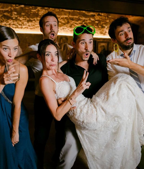 Un grupo de personas, una de ellas con vestido de novia y zapatillas deportivas, posa de forma divertida en un interior. Algunas llevan ropa informal y una lleva gafas de sol de color verde neón en la cabeza.