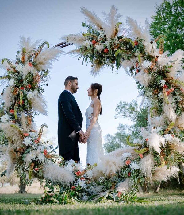 Una pareja se toma de la mano dentro de una gran corona de flores al aire libre, rodeada de vegetación.