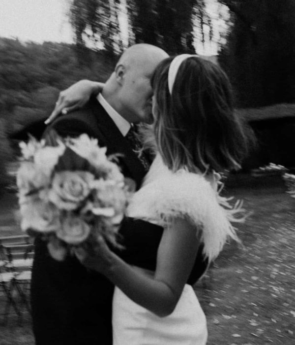 Una pareja se abraza y se besa al aire libre, la mujer sostiene un ramo de flores. Hay un fondo borroso de sillas dispuestas en hileras sobre una zona de césped.