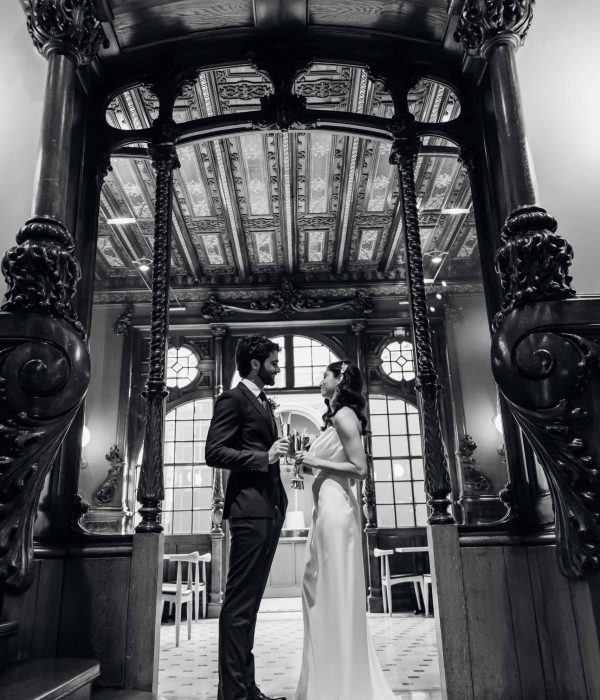 Una pareja vestida formal se encuentra frente a frente, tomados de la mano, en un interior histórico adornado con columnas de madera decorativas y detalles tallados en el techo.