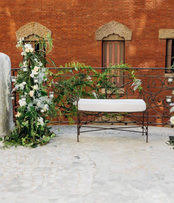 Un banco de hierro forjado con cojines blancos está flanqueado por altos arreglos florales en una terraza de baldosas con paredes de ladrillo en el fondo.