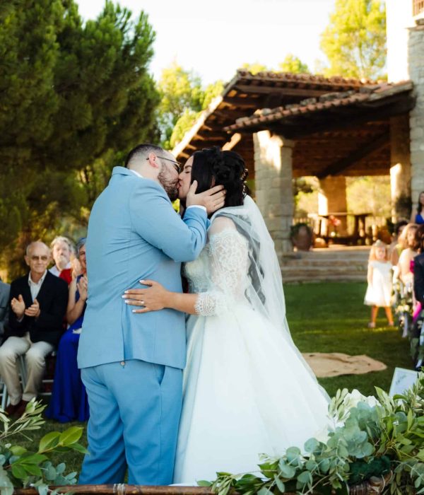 Una novia y un novio se besan al aire libre frente a los invitados que aplauden. La escena incluye vegetación y un edificio de piedra al fondo.