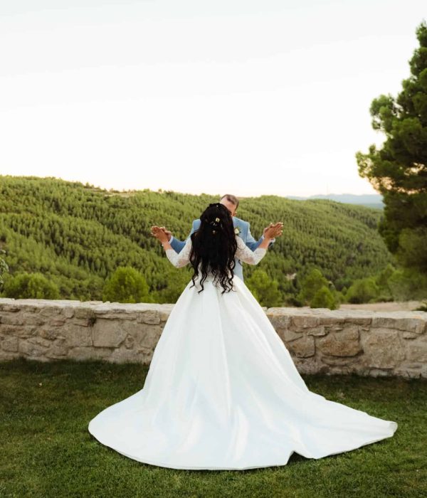 Una novia con un vestido de novia blanco se encuentra al aire libre, frente a una vista de exuberantes colinas verdes y árboles, con los brazos levantados.