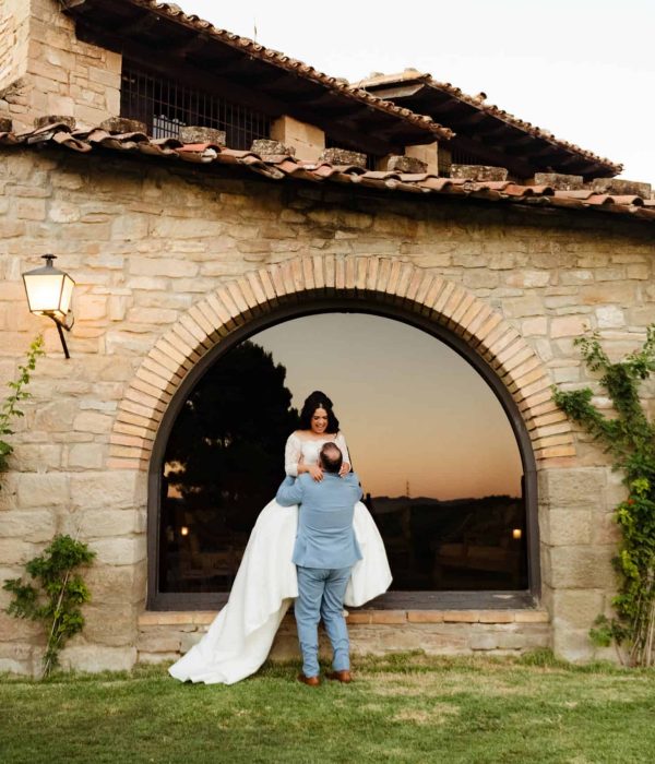 Una persona con vestido de novia es levantada por otra persona frente a un edificio de piedra con una gran ventana arqueada.
