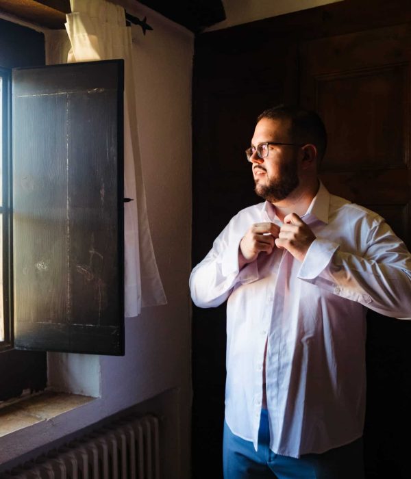 Un hombre está de pie junto a una ventana, abrochándose la camisa, mientras la luz del sol entra a raudales.