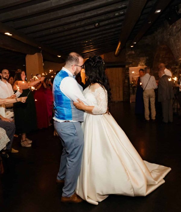 Una pareja baila en la recepción de una boda rodeada de invitados vestidos de manera formal y sosteniendo bengalas en una habitación poco iluminada.
