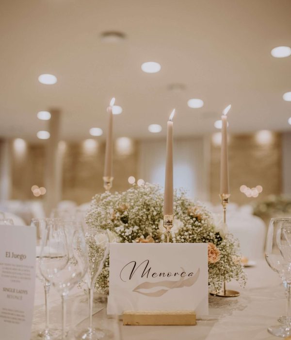 Elegante mesa de comedor con velas altas, un centro de mesa floral y una tarjeta "Menorca", ubicada en una habitación suavemente iluminada.