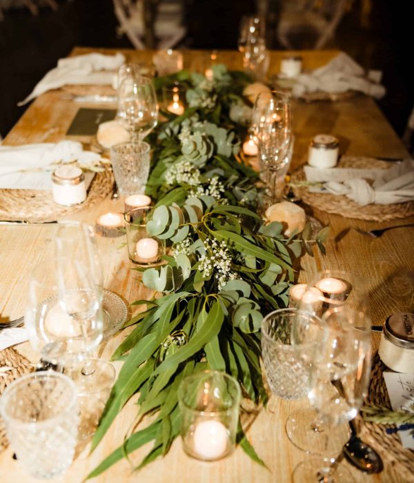 Una mesa de madera está preparada para una cena con vegetación y flores blancas como centro de mesa, rodeada de velas, cristalería y servilletas blancas.