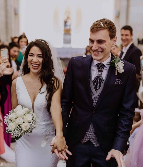 Una novia y un novio sonríen mientras caminan hacia el altar, rodeados de invitados que aplauden. La novia sostiene un ramo de rosas blancas.