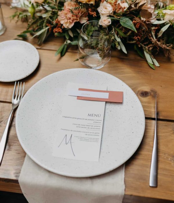 Una mesa de madera con un plato moteado, una tarjeta de menú, una tarjeta con nombres, un tenedor, un cuchillo, un vaso y un centro de mesa floral.
