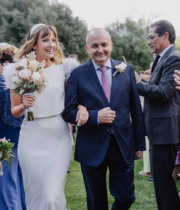Una novia con un vestido blanco y un ramo de flores en la mano camina del brazo con un hombre mayor con traje por un pasillo al aire libre, rodeada de invitados que aplauden.