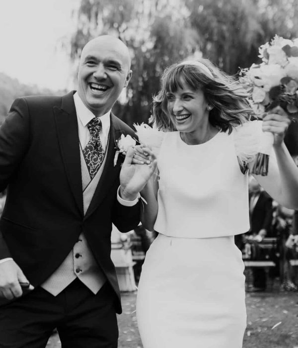 Una pareja vestida de boda, alegremente tomadas de la mano y con flores, rodeadas de una multitud que los anima al aire libre.