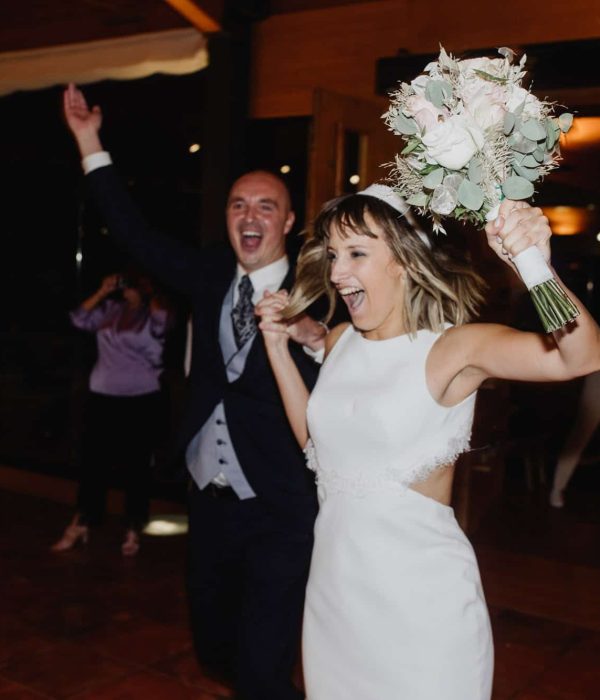 Una novia y un novio entran alegremente en un salón de recepción. La novia sostiene un ramo de flores y levanta la mano emocionada. Ambos sonríen y el ambiente es festivo.