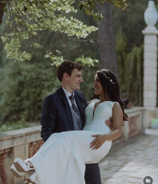 Un novio con traje lleva a una novia con un vestido blanco al aire libre, con árboles y barandilla de piedra al fondo.