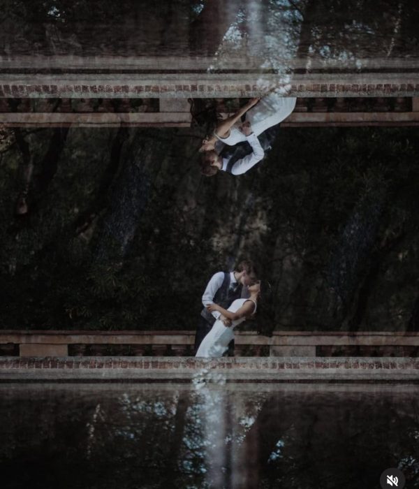 Una pareja vestida formalmente se abraza en un puente, reflejada en el agua de abajo.