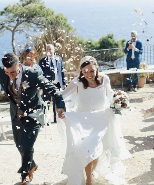 Una novia y un novio caminan alegremente de la mano por un pasillo al aire libre mientras los invitados arrojan pétalos de flores. El mar y los árboles se ven al fondo.
