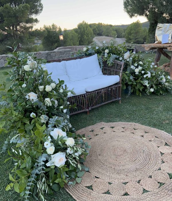 Un banco de mimbre blanco con cojines rodeado de follaje verde y flores blancas en una zona de césped, junto a una alfombra circular tejida.