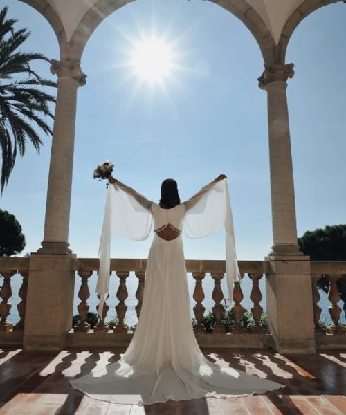 Una persona con un vestido largo blanco se encuentra de pie en una terraza con los brazos en alto y sosteniendo un ramo de flores. La escena muestra arcos, una palmera y la luz del sol contra un cielo despejado.