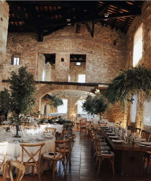 Un comedor rústico con paredes de piedra, ventanas arqueadas y vigas de madera. Las mesas están preparadas para un evento formal con manteles blancos, cristalería y centros de mesa con flores verdes.