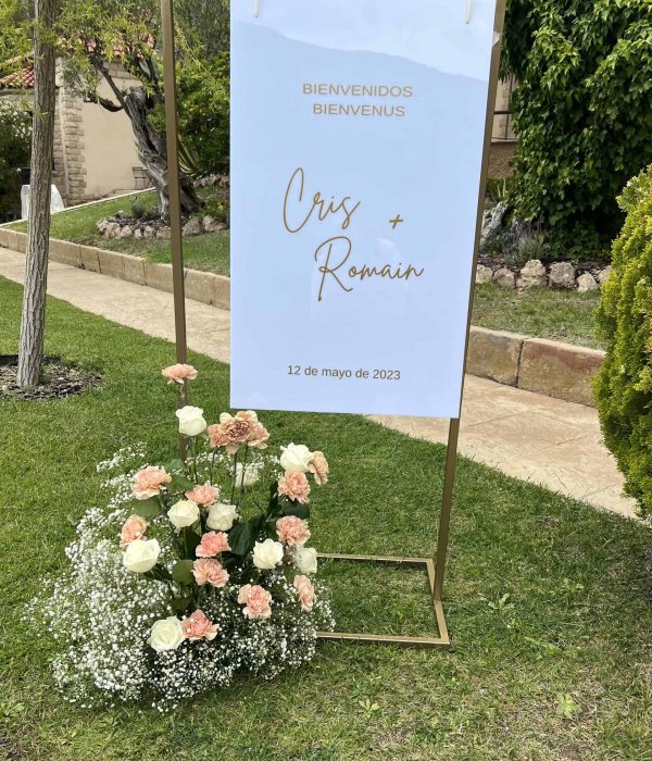 Cartel de bienvenida de boda para Cris y Romain con fecha del 12 de mayo de 2023, sobre soporte con flores blancas y durazno en la base, colocado sobre césped.