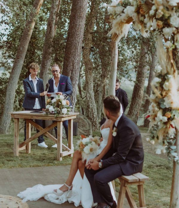 Una pareja se sienta bajo un arco de flores durante una ceremonia nupcial al aire libre, mientras dos personas están de pie detrás de una mesa de madera sosteniendo papeles. Los árboles forman el fondo.