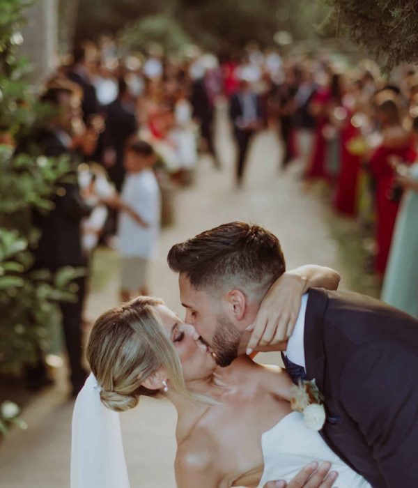 Una novia y un novio comparten un beso en un camino bordeado de árboles con una multitud de personas en el fondo.