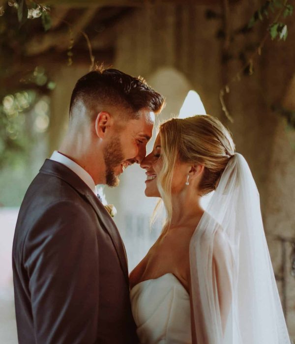 Una novia y un novio se miran uno al otro, sonriendo suavemente, en un entorno al aire libre iluminado por el sol, con arcos y vegetación.