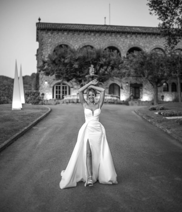 Una persona con un vestido sin tirantes posa con los brazos en alto en un camino de entrada frente a un gran edificio de piedra.