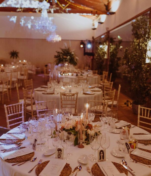 Elegante salón de banquetes con mesas redondas preparadas para una cena formal. Las mesas cuentan con manteles blancos, centros de mesa florales y velas encendidas. Candelabros de cristal cuelgan del techo de madera.