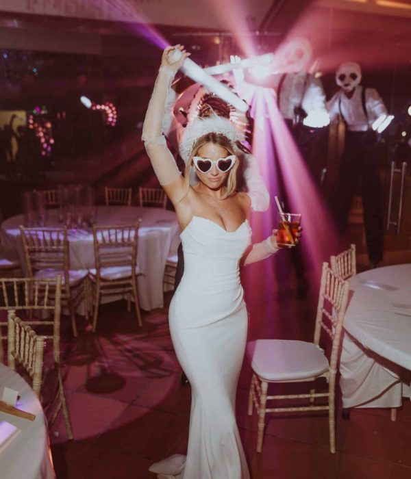 Una persona con un vestido de novia y gafas de sol en forma de corazón baila enérgicamente en una fiesta, sosteniendo una bebida en una mano y un palo brillante en la otra, con los asistentes a la fiesta en el fondo.