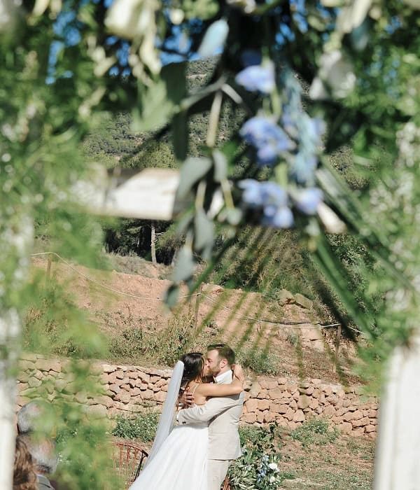 Una pareja se besa bajo un arco floral en una ceremonia de boda al aire libre, con vegetación y flores azules en primer plano.