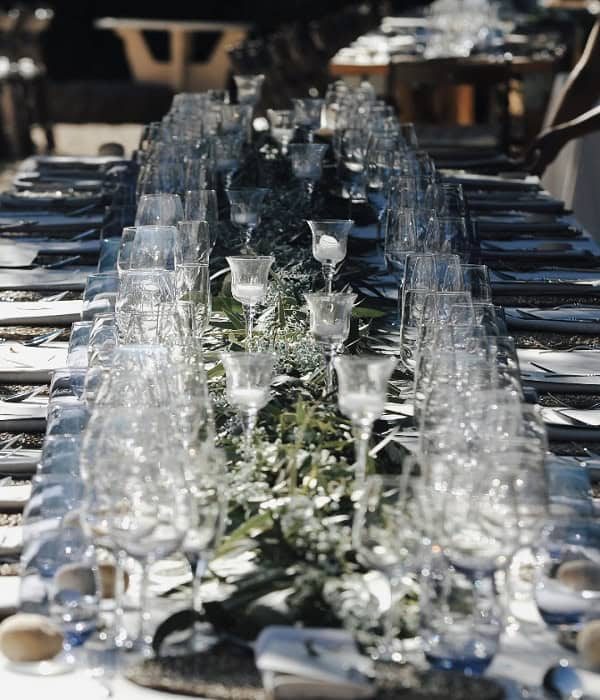 Una mesa de comedor larga está elegantemente dispuesta con cristalería, servilletas blancas y vegetación decorativa que recorre el centro.