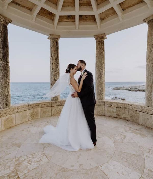 Una pareja se abraza bajo un cenador de piedra junto al mar, con la novia con un vestido blanco y el novio con un traje negro.