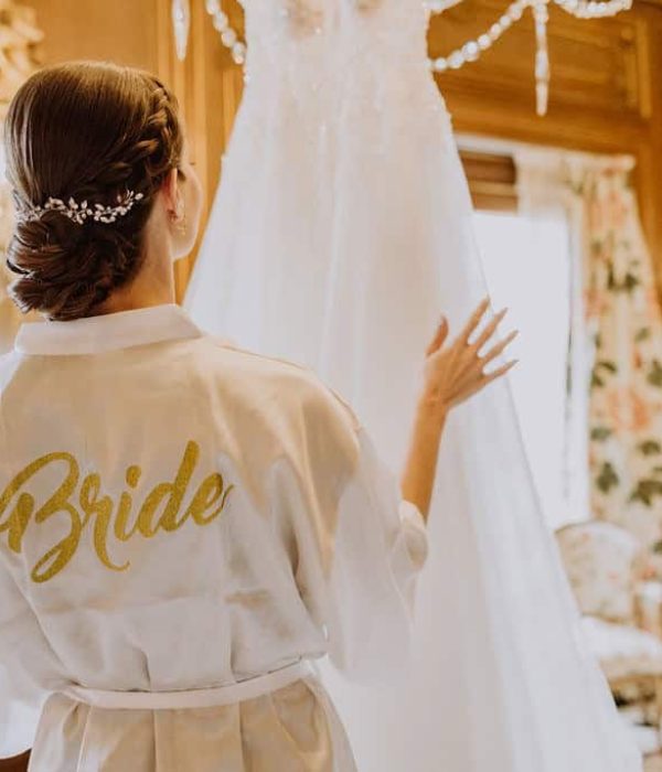 Una mujer que viste una túnica con una etiqueta que dice "Novia" mira un vestido de novia colgado en una habitación ornamentada con paredes revestidas de madera y una lámpara de araña.