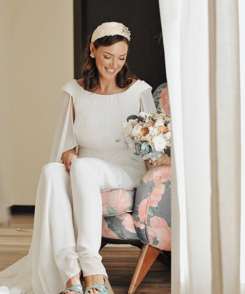 Una mujer con un vestido blanco está sentada en una silla con flores, sosteniendo un ramo y usando zapatos azules.