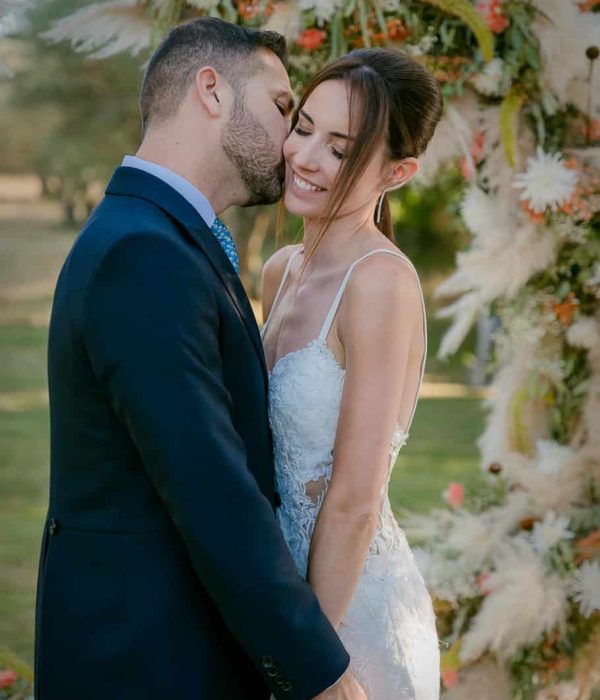 Una pareja de novios se abraza bajo un arco de flores al aire libre. El hombre le susurra algo al oído a la mujer mientras ella sonríe.